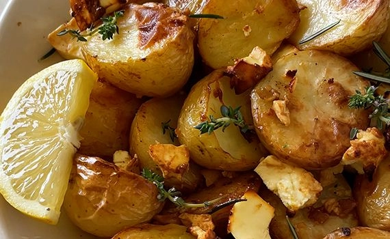 A close up of a dish of lemon feta roasted potatoes with herbs and a garnish of lemon slice