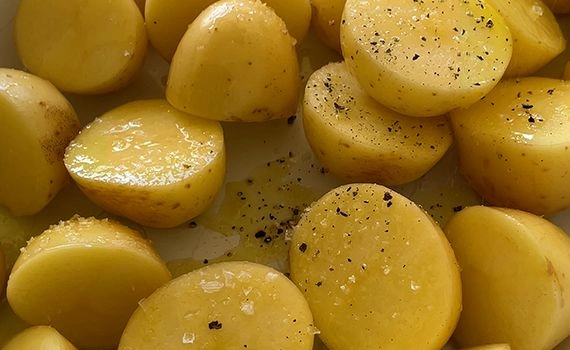 Olive oil, salt and pepper added to cut baby potatoes in a tray.