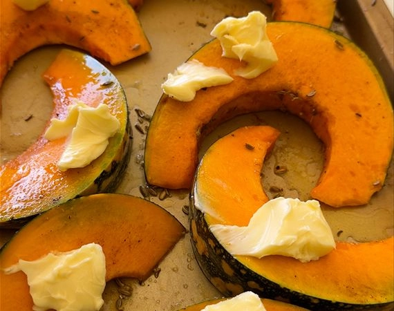 Pumpkin wedges on a roasting tray with oil, spices, seasoning and dollops of butter.